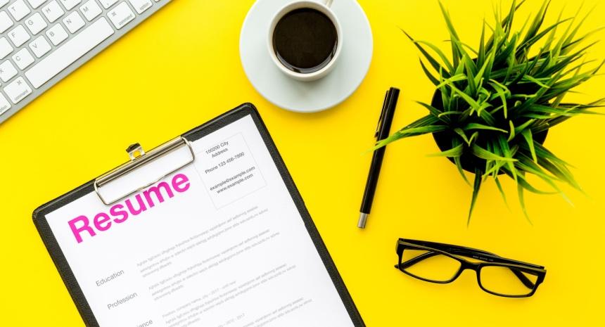 image of resume on yellow desk along with pen, keyboard, cup of coffee, desk plant, and reading glasses